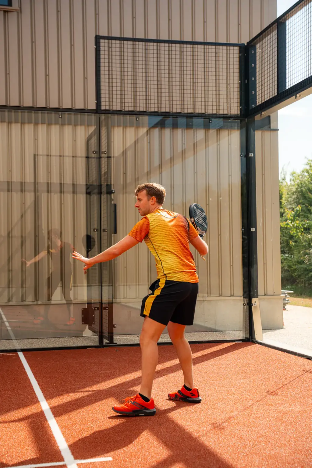 Joueur amorçant un revers le long de la paroi vitrée d'un court de padel extérieur