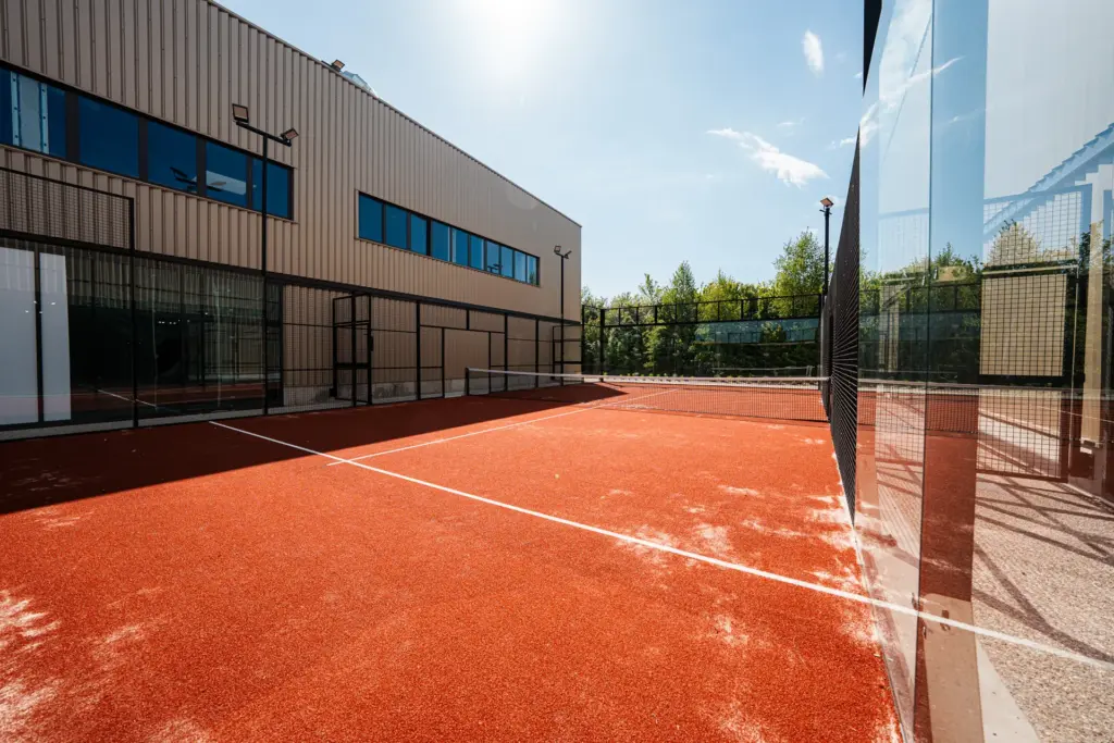Terrain de padel extérieur longeant la façade vitrée du club, arbres en arrière-plan près de Rouen