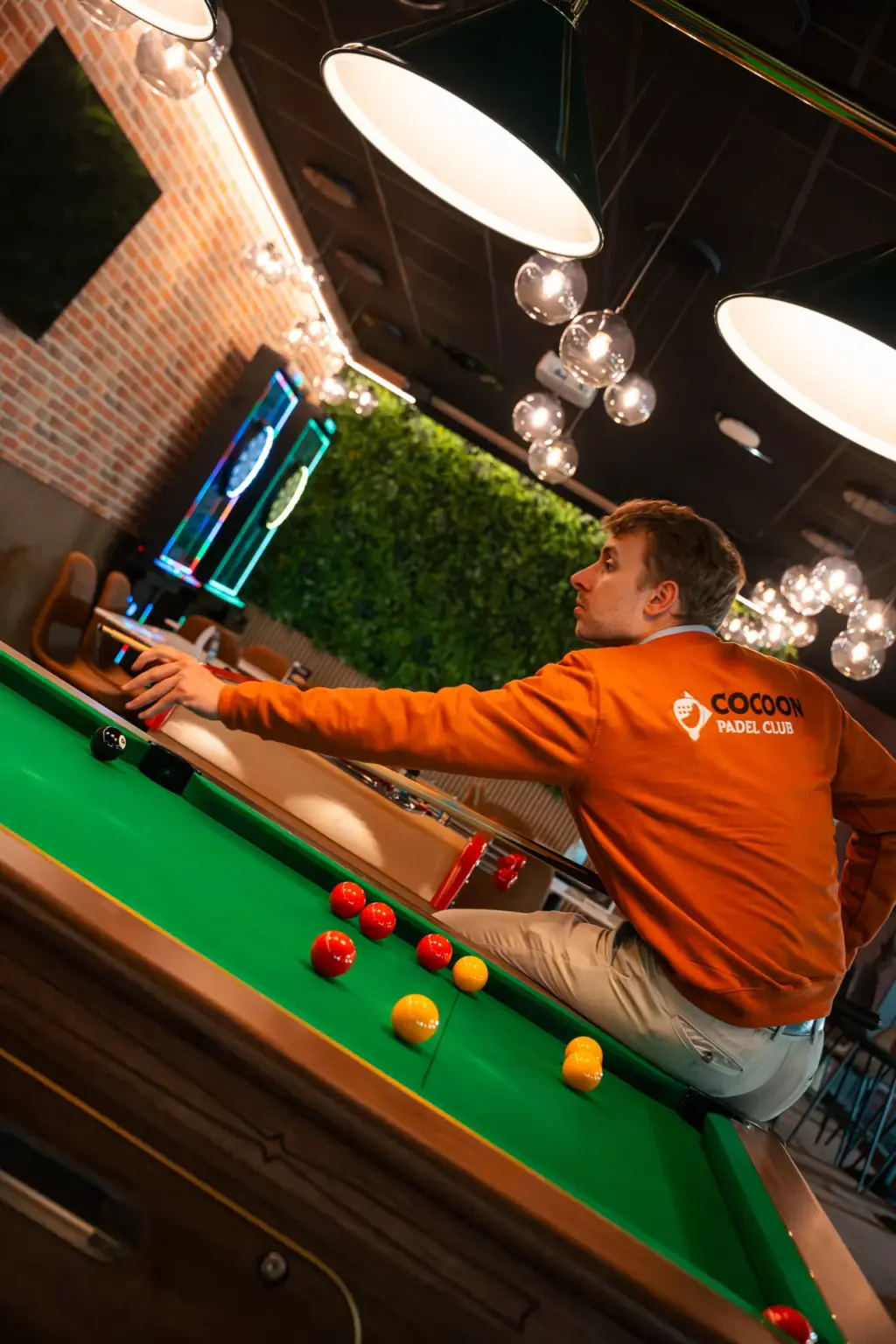 Joueur en sweat Cocoon Padel Club au billard devant le mur végétal du lounge, ambiance conviviale